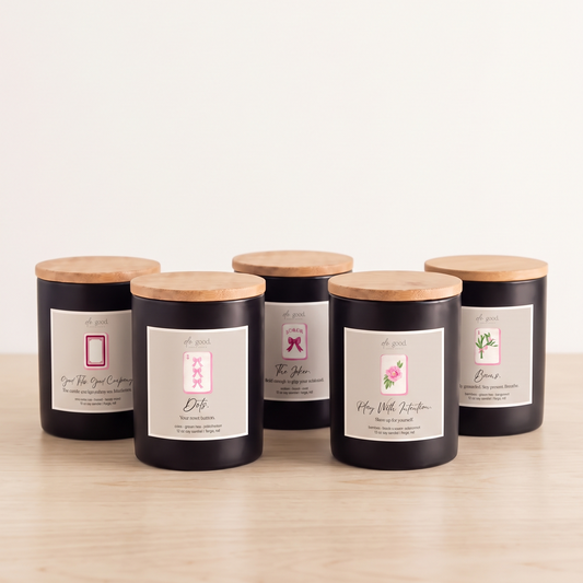 Five black glass jar candles with light wood lids and white labels are arranged on a light wood surface against a plain white background.