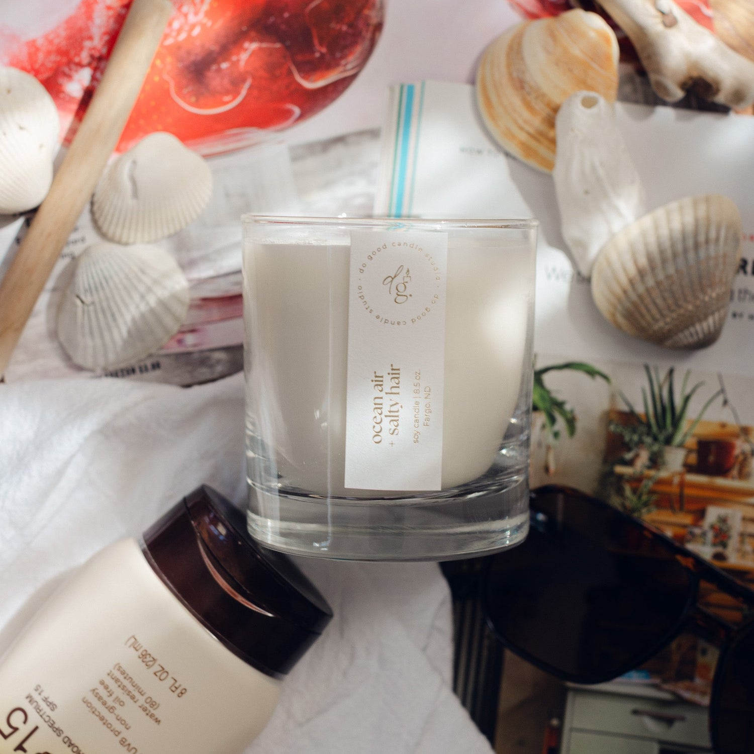 flatlay of ocean air + salty hair candle with shells, sunscreen, magazines around it