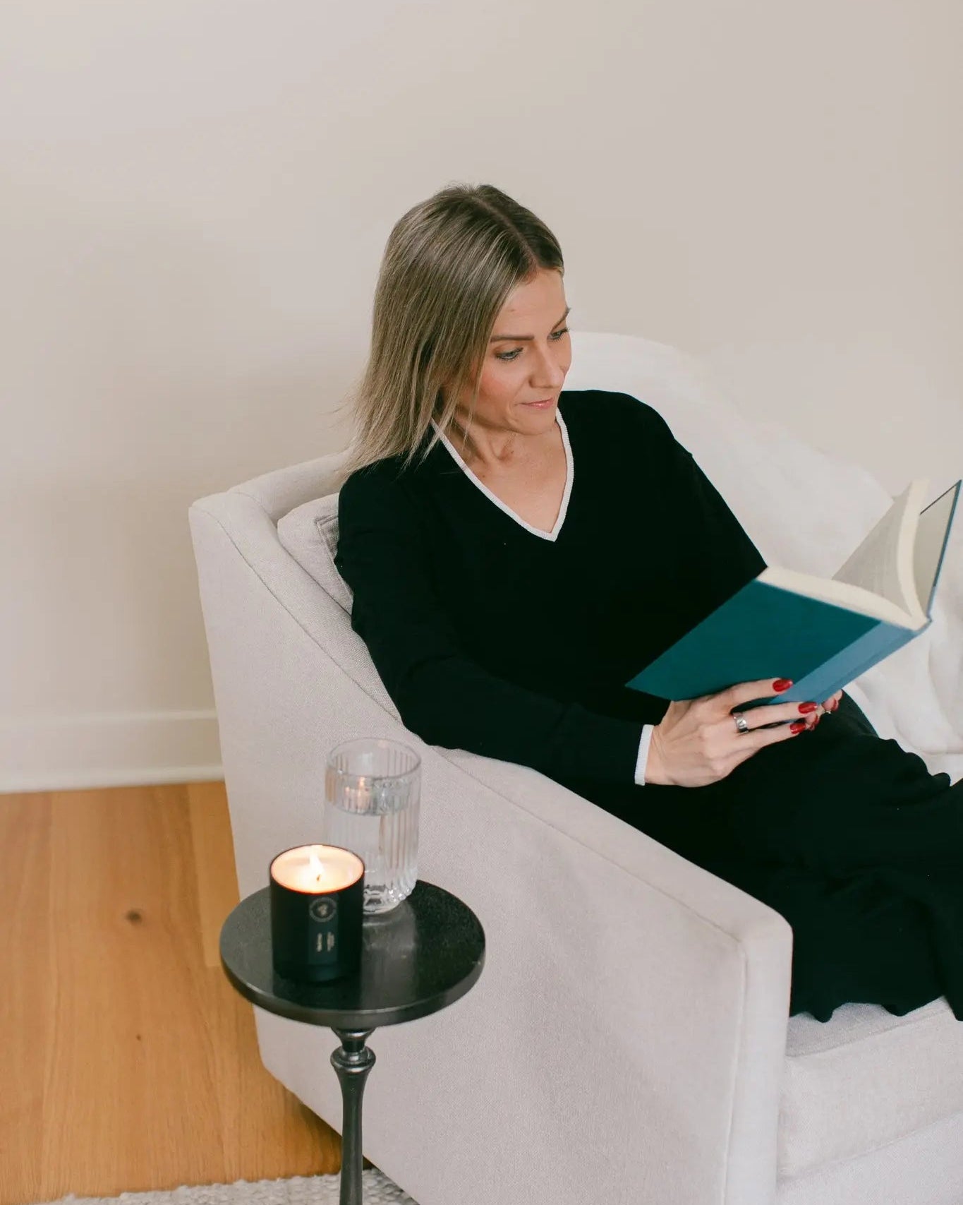 Woman reading a book in a white armchair with a candle and glass of water on a small table.