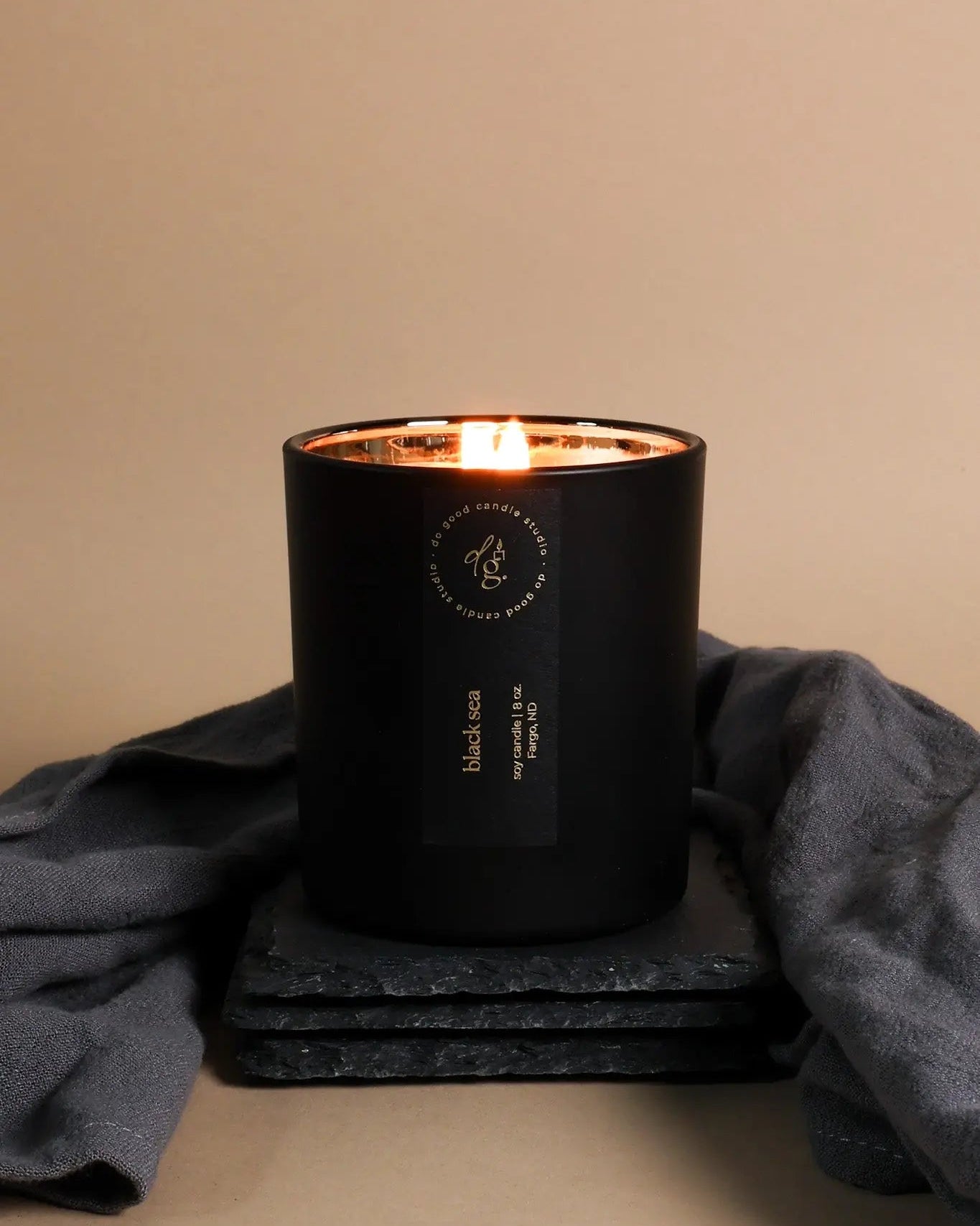 soy candle in matte black vessel with a lit wick on a dark surface with a beige background