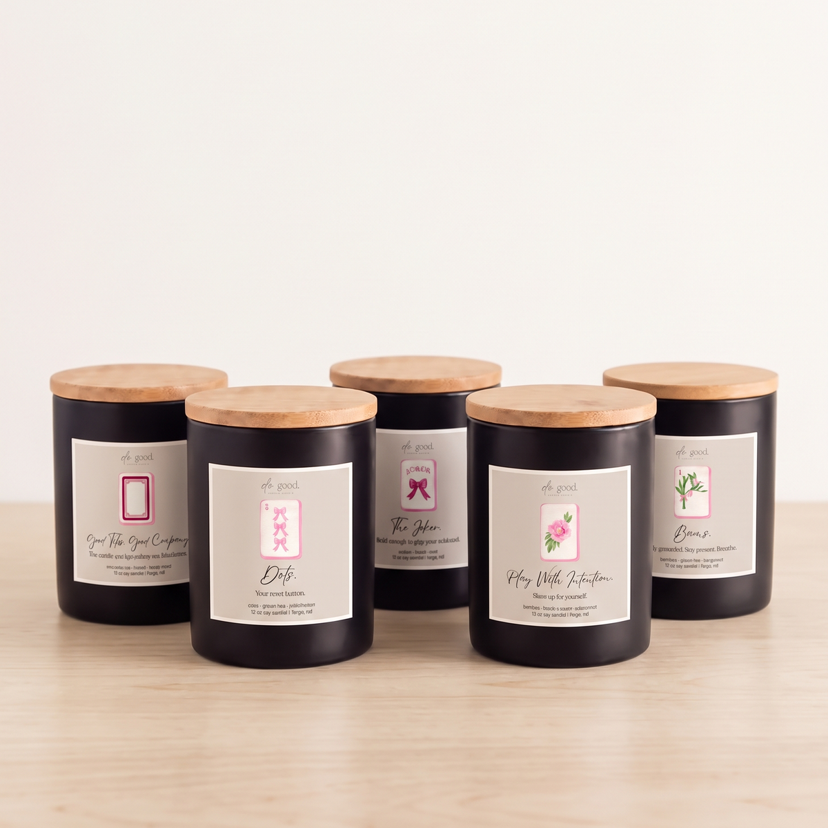 Five black glass jar candles with light wood lids and white labels are arranged on a light wood surface against a plain white background.