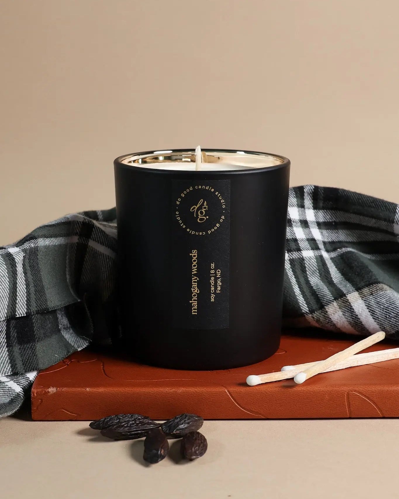 Mahogany Woods candle styled on a leather journal with a plaid cloth, matches and tonka beans