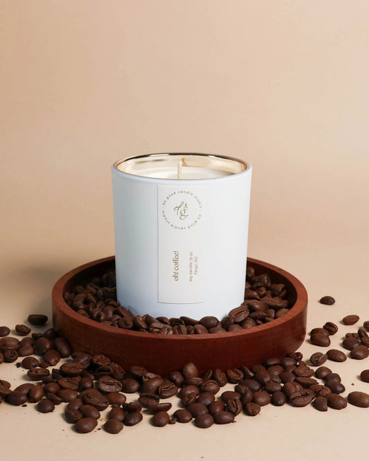Oh! Coffee! candle styled on a beige backdrop with a wood coaster full of coffee beans