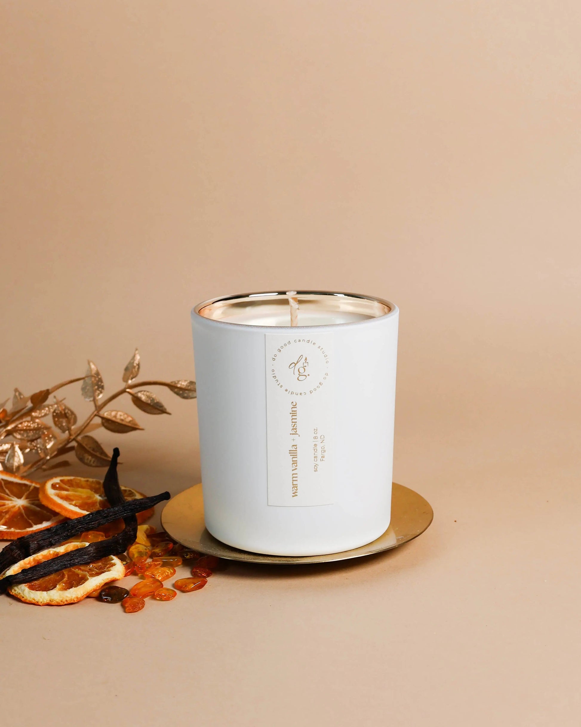 Warm Vanilla + Jasmine candle styled o a beige backdrop with a silver coaster, gilded branches, vanilla beans, amber stones and citrus slices