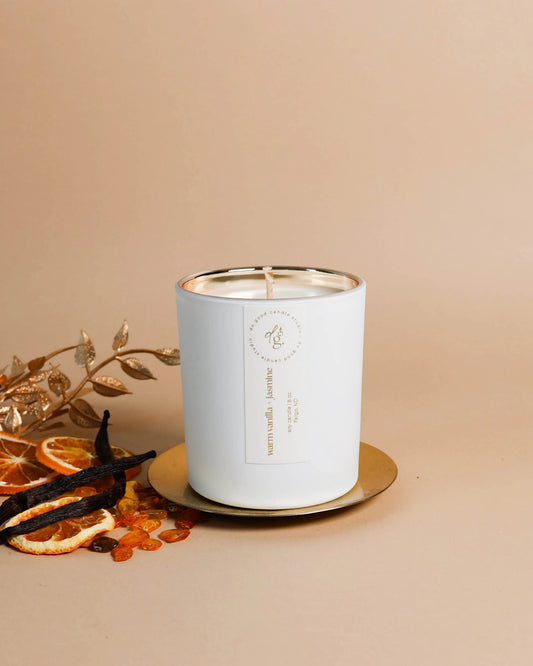 Warm Vanilla + Jasmine candle styled o a beige backdrop with a silver coaster, gilded branches, vanilla beans, amber stones and citrus slices